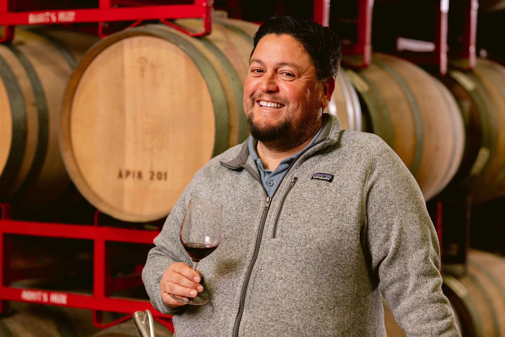 Macario Montoya, winemaker at Roots Run Deep Winery, smiling in the barrel room with a glass of red wine